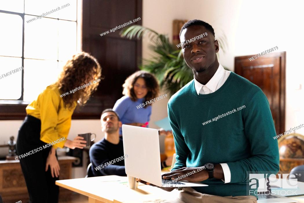 Stock Photo: Portrait of a confident man in office using laptop with colleagues in background.