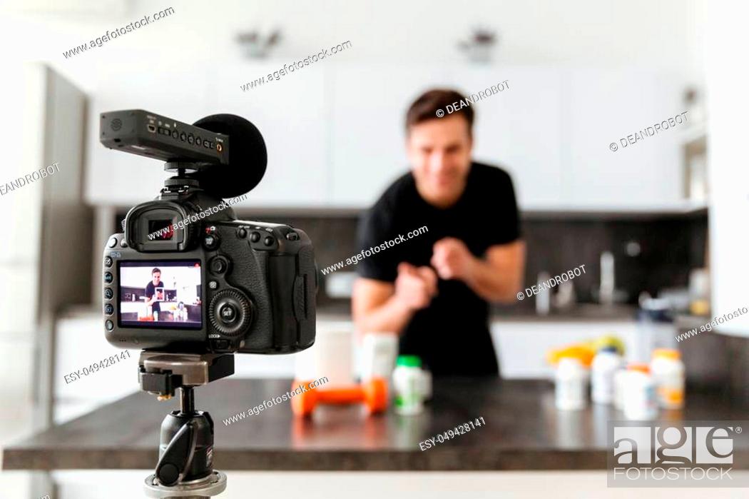 Stock Photo: Happy young man filming his video blog episode about healthy food additives while standing at the kitchen table and pointing finger at camera.