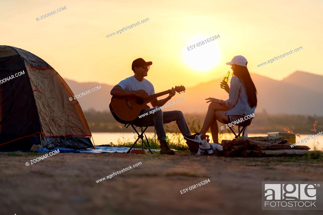 Stock Photo: Young adult Asian couple playing guitar and drinking beer beside their tent campsite while sunset. Camping trip with dog outdoor activity campsite concept.