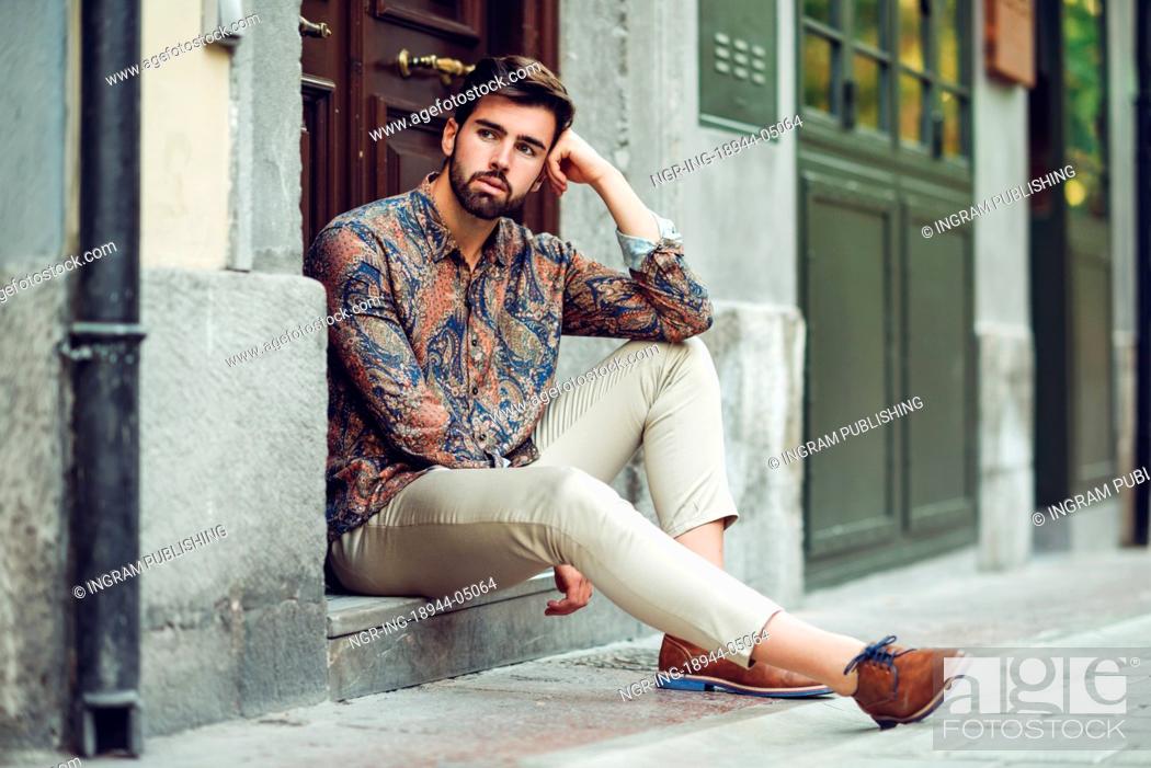 Stock Photo: Young bearded man, model of fashion, sitting in an urban step wearing casual clothes. Guy with beard and modern hairstyle looking away in the street.