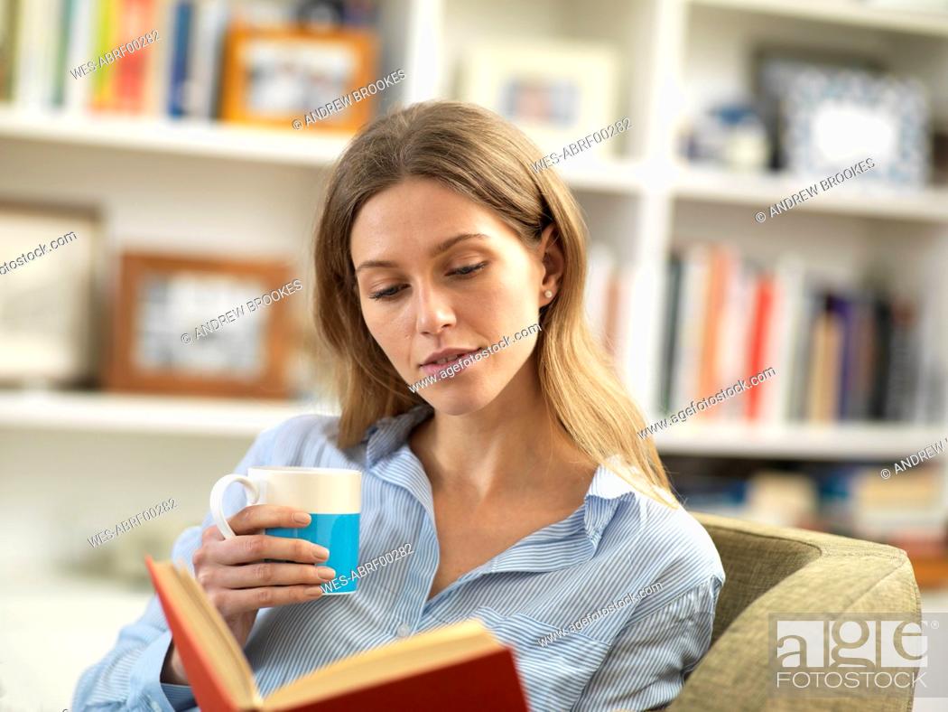 Photo de stock: Young woman with a hot drink relaxing at home reading a book.