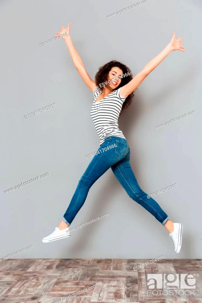 Stock Photo: Energetic woman 20s in striped t-shirt and jeans, jumping with hands throwing up in air over grey background.