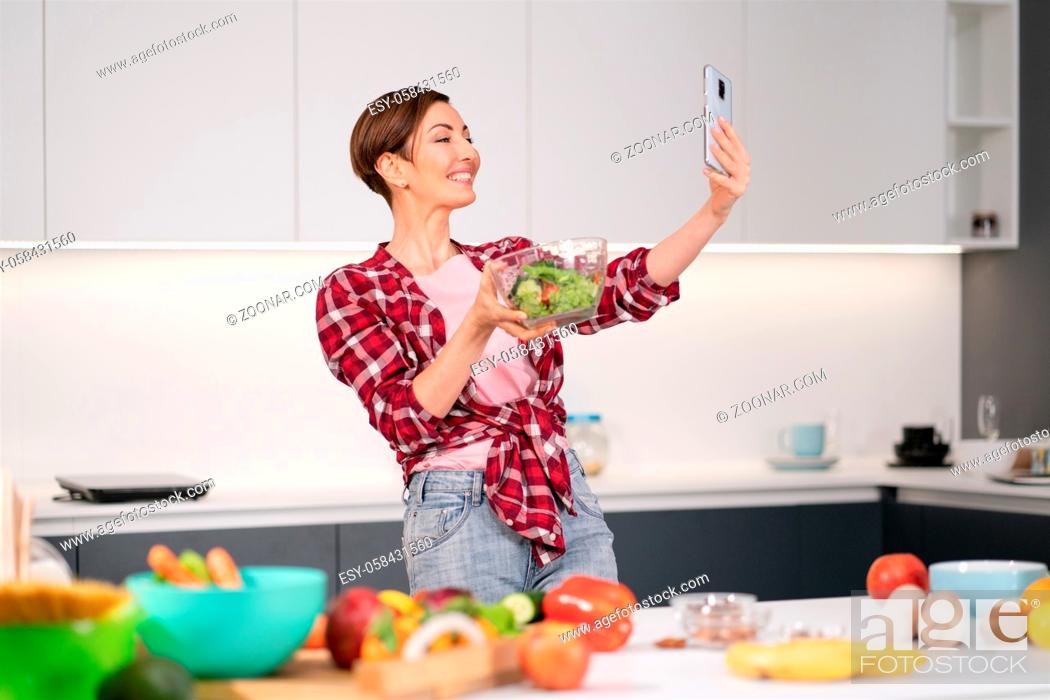 Stock Photo: Pretty woman happy taking selfie using her smartphone while cooking fresh salad wearing a plaid shirt with a bob hair style.