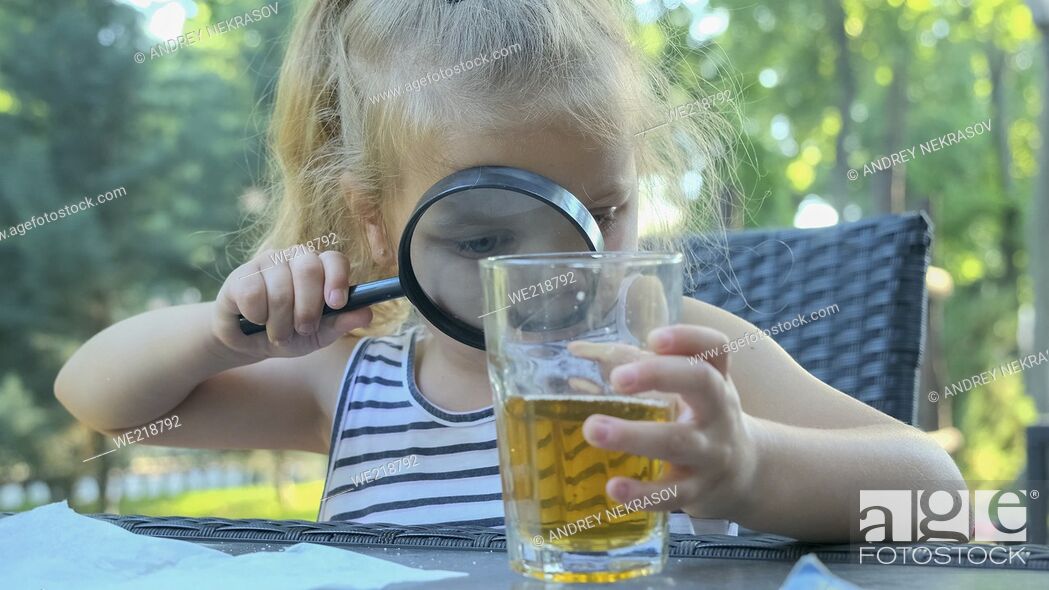 Stock Photo: Cute little girl carefully looks into the lens at the juice in the glass. Close-up of blonde girl studying her juice in glass while looking at her through.