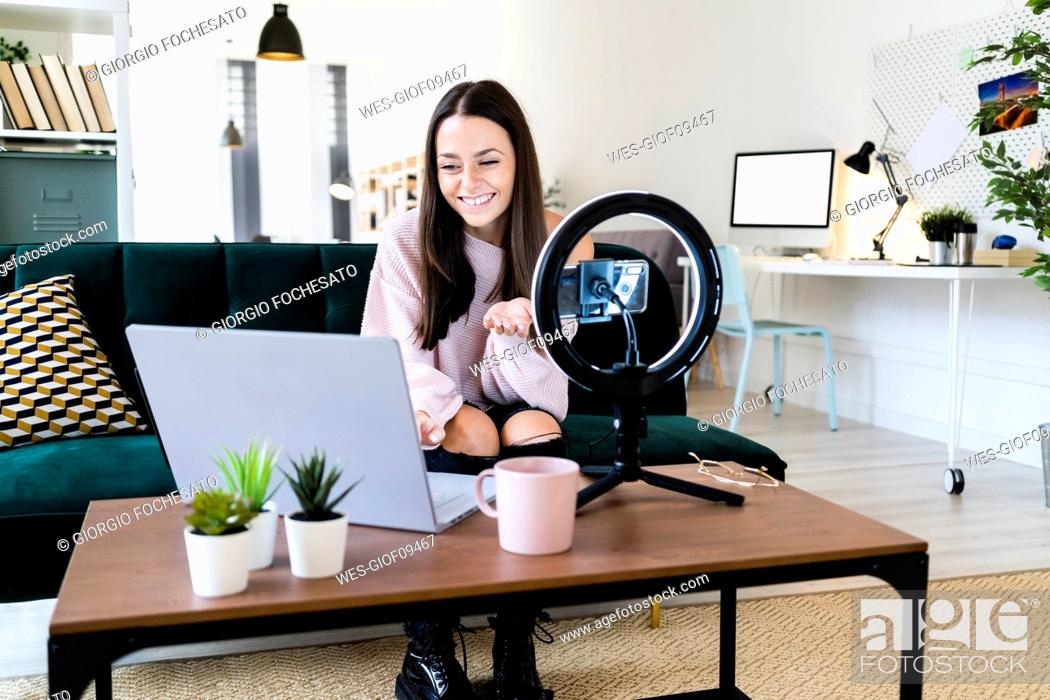 Stock Photo: Smiling beautiful female vlogger using laptop while filming through smart phone at loft apartment.