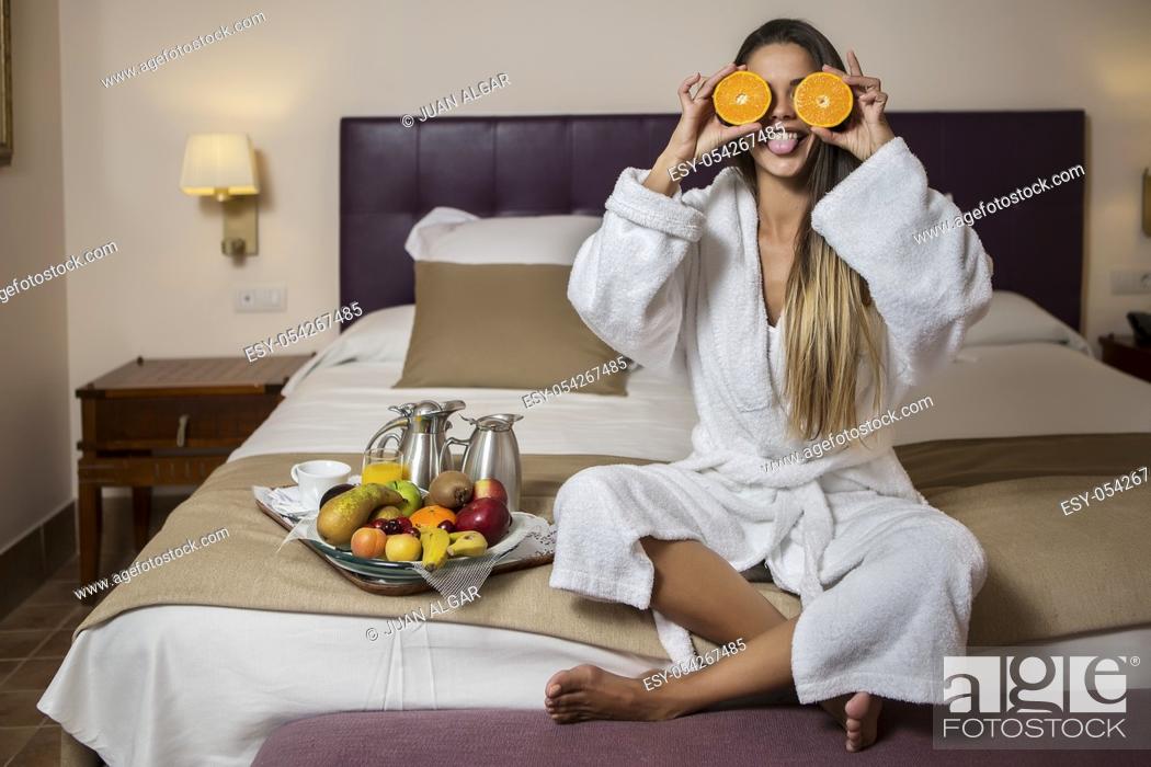 Stock Photo: Young pretty woman in hotel morning gown sitting on bed with slices of orange near eyes and showing tongue with plate of fresh fruits placed near.