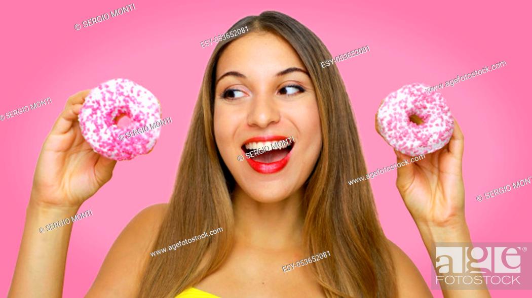 Imagen: Happy cheerful excited young woman holding two donuts in her hands looking to the side on pink background.