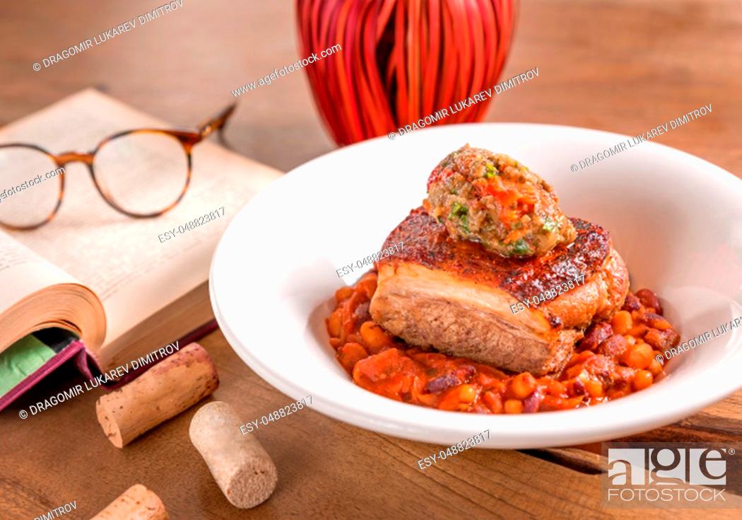 Photo de stock: Pork belly with bean stew and mashed vegetables, served in a white plate, decorated with book and glasses on a wooden table.