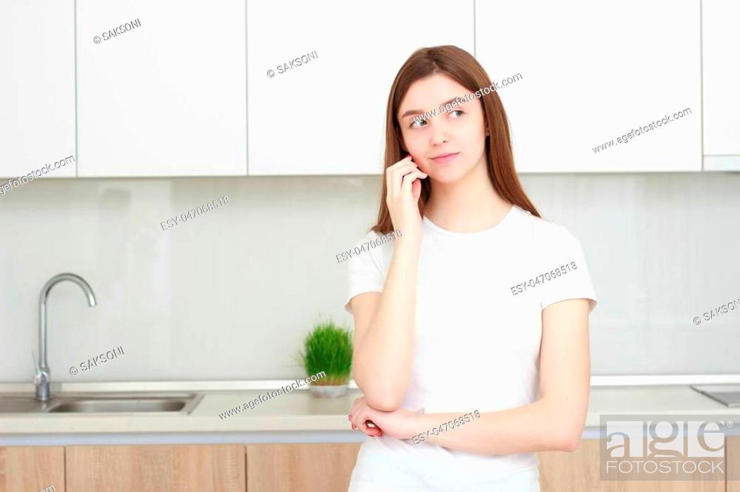 Stock Photo: Pretty young woman In a white T shirt standing in kitchen and smiling. Real interior.