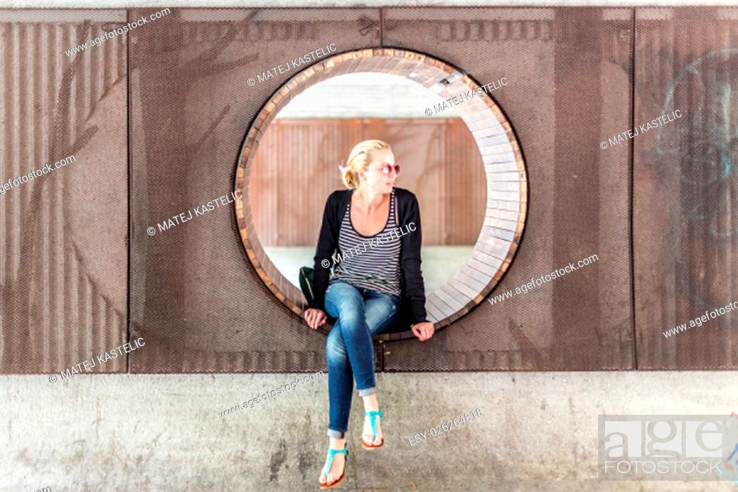Stock Photo: Casually dressed young urban lady wearing red sunglasses sitting on contemporary circular wooden bench in city park.