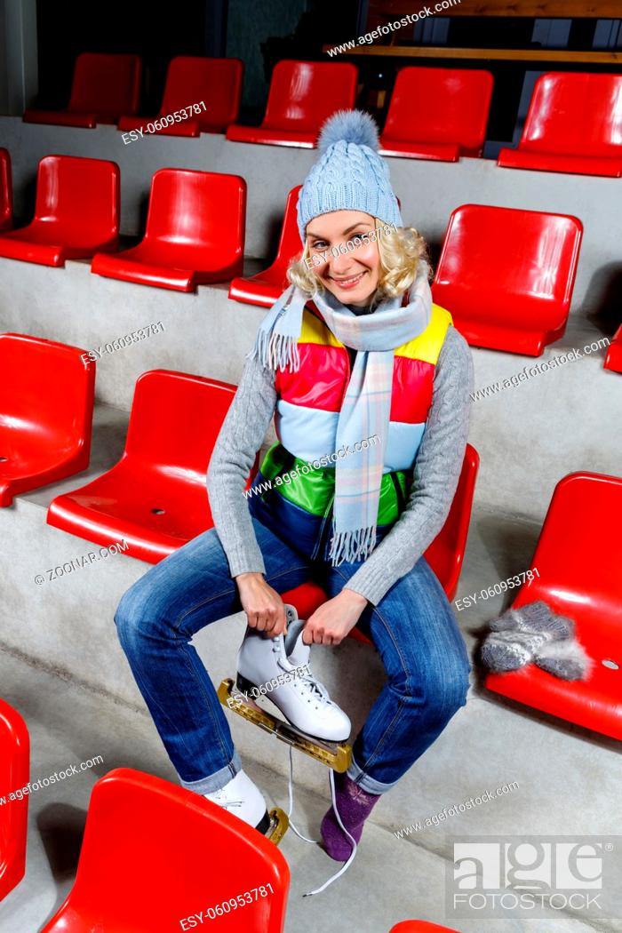 Stock Photo: Beautiful blond young woman in winter clothes sitting on red tribune and putting on white skates. Girl getting ready for skating on ice rink.