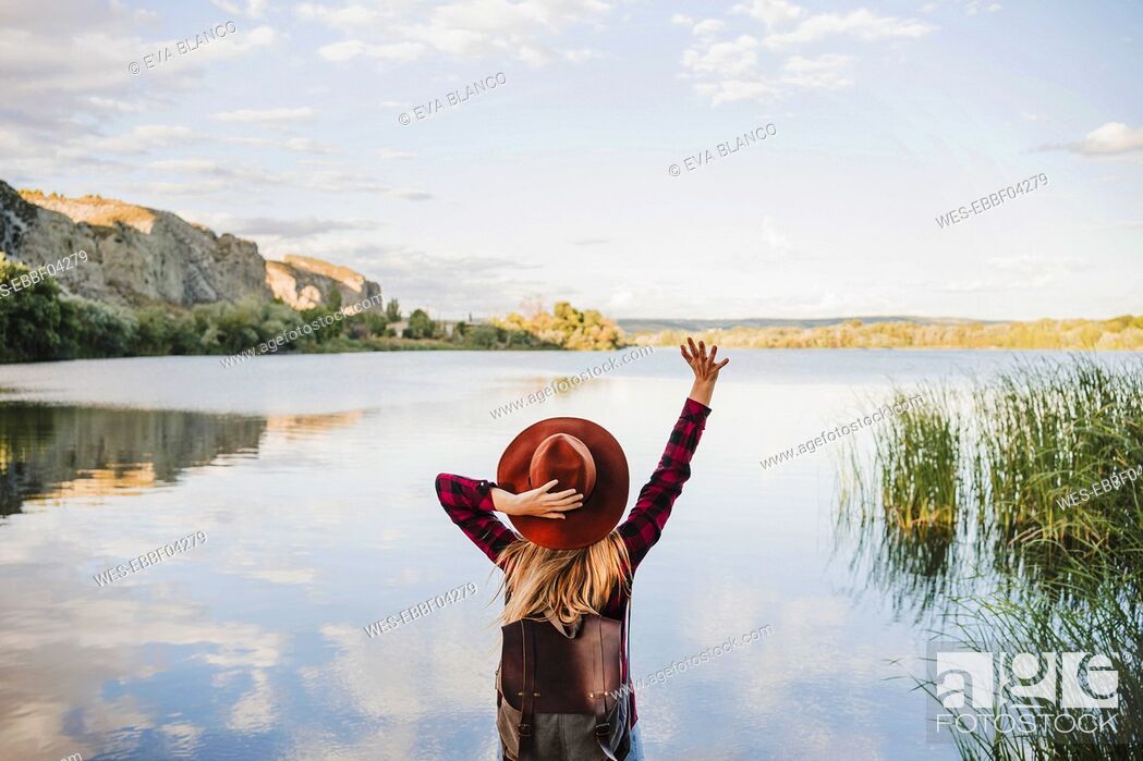 Stock Photo: Young blond woman holding hat while standing with hand raised at lakeshore.