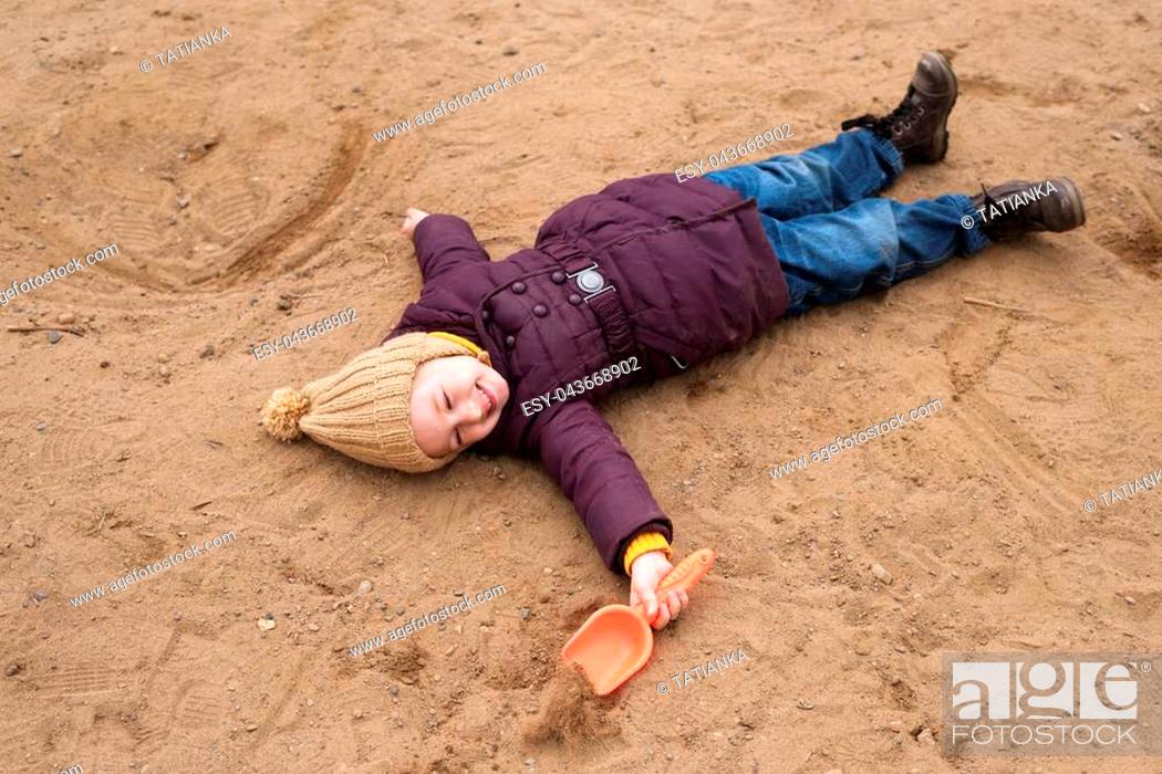 Stock Photo: Little girl in a violet coat and a beige woolen hat lying on the sand in a sand-box with a plastic shovel in her hand.