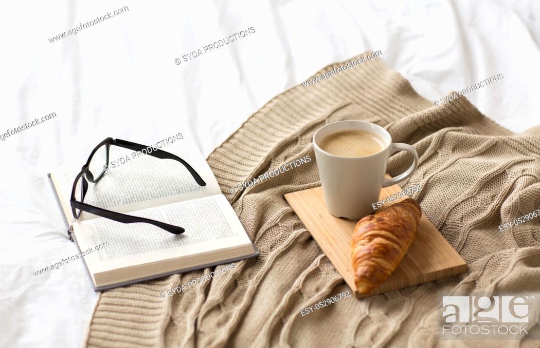 Imagen: coffee, croissant, blanket and book on bed at home.