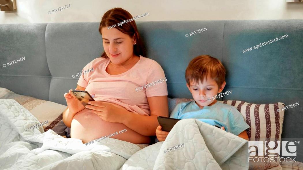 Stock Photo: Young pregnant mother with little son lying in bed and using smartphone and tablet computer before going to sleep at night.