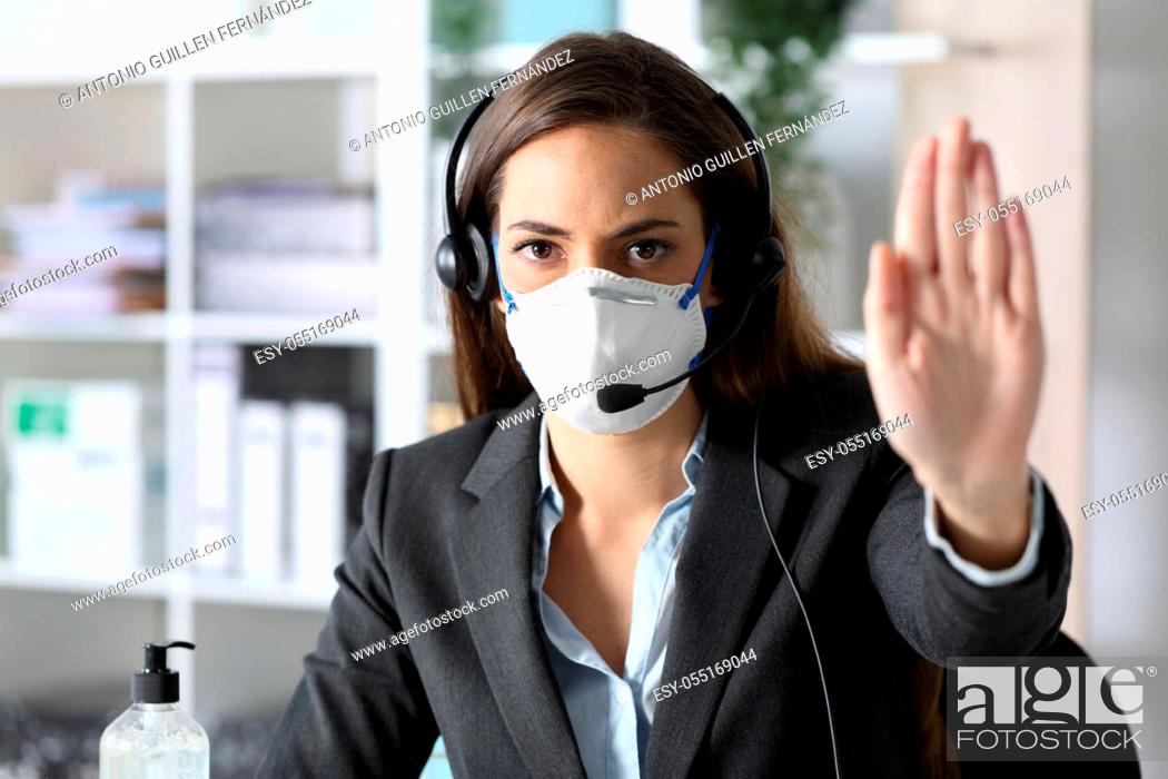 Imagen: Serious telemarketer woman gesturing stop avoiding covid-19 with mask looking at camera sitting at the office.