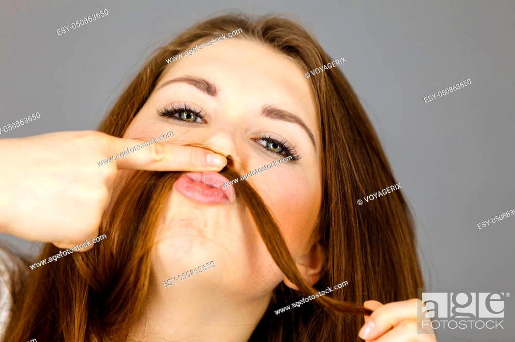 Stock Photo: Woman having fun with her long brown hair making moustache being happy about hairdo condition, studio shot grey background.