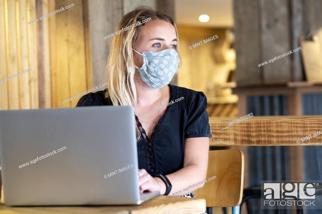 Stock Photo: Young blond woman wearing blue face mask, sitting at table, using laptop computer.