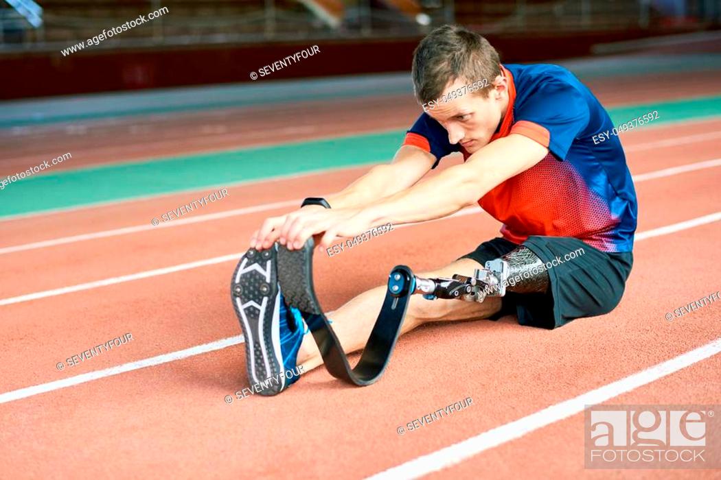 Stock Photo: Portrait of young amputee sportsman warming up before running practice in modern gym stretching muscles sitting on track, copy space.