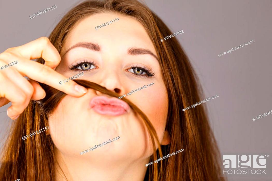 Stock Photo: Woman having fun with her long brown hair making moustache being happy about hairdo condition, studio shot grey background.