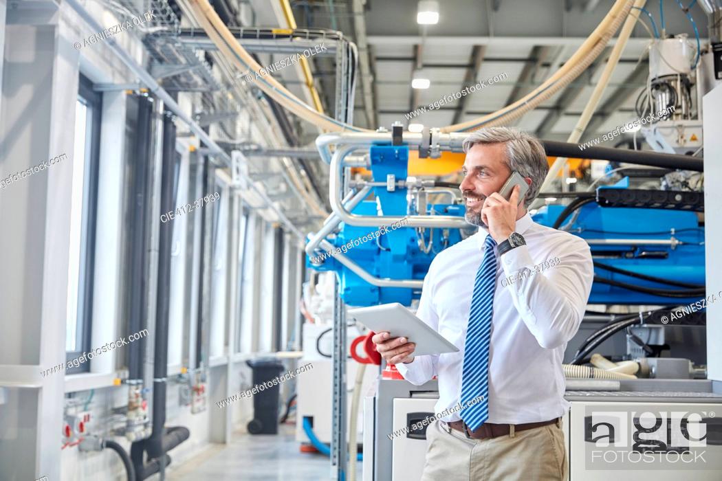 Stock Photo: Male supervisor with digital tablet talking on cell phone next to plastic industrial molder machinery in factory.