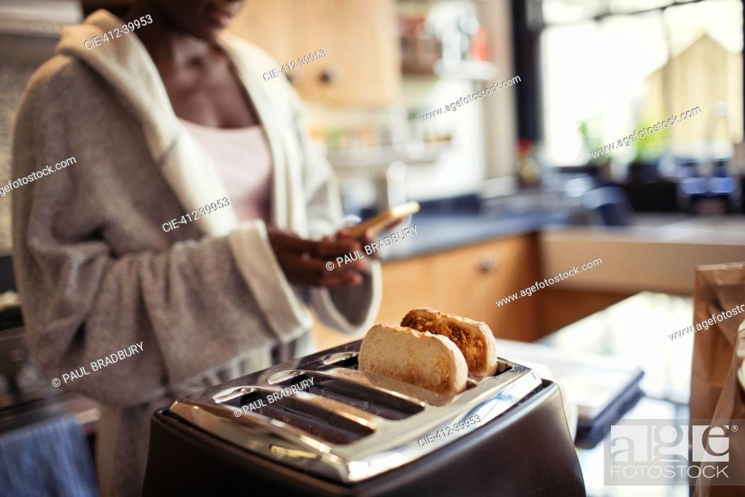 Stock Photo: Woman texting with smart phone, toasting bread in toaster in kitchen.