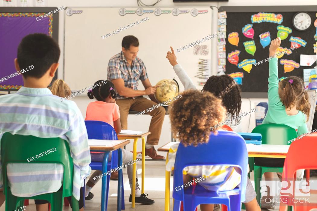 Stock Photo: Front view of schoolteacher learning at his pupils the earth globe in classroom at school with school kids looking at him and raising hand in foreground.