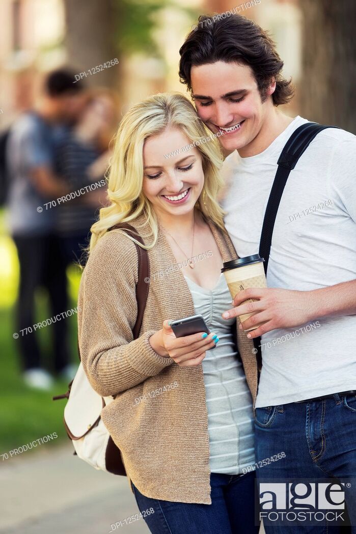 Stock Photo: A young couple standing together and checking social media on a smart phone while walking through a university campus; Edmonton, Alberta, Canada.