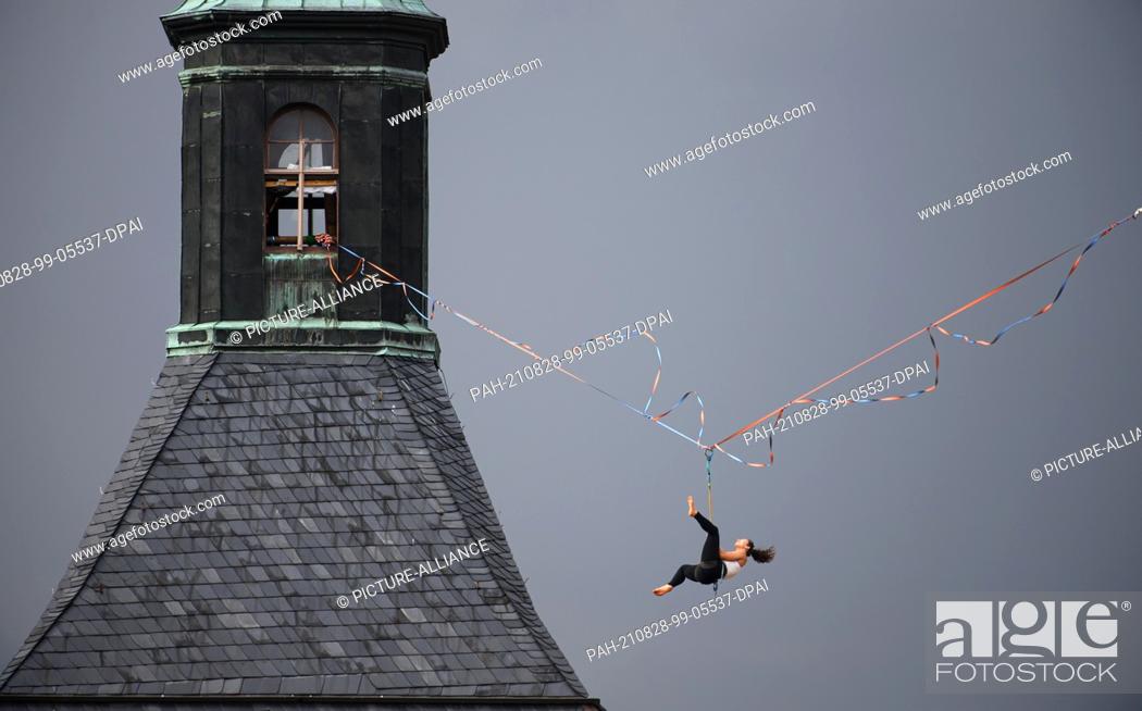 Stock Photo: 28 August 2021, Saxony, Hohnstein: A slackline runner hangs on a rope between the castle and the town church of the town in Saxon Switzerland at the Highline -.