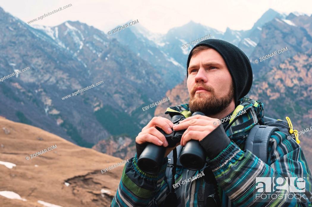 Stock Photo: A hipster man with a beard in a hat, a jacket, and a backpack in the caucasian mountains holds binoculars, adventure, tourism, tracking.