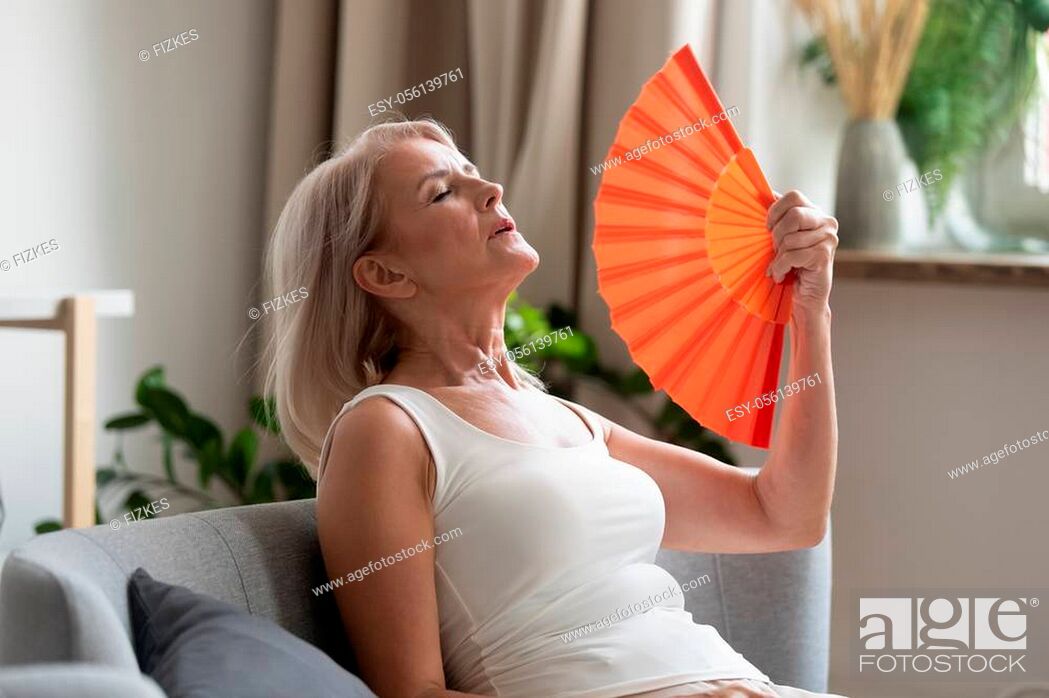 Photo de stock: Stressed annoyed old senior woman using waving fan suffer from overheating, summer heat health hormone problem, no air conditioner at home sit on sofa feel.