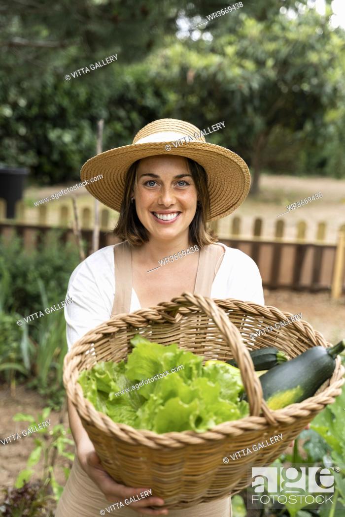 Stock Photo: Smiling young woman holding wicker basket with vegetables in yard during curfew.