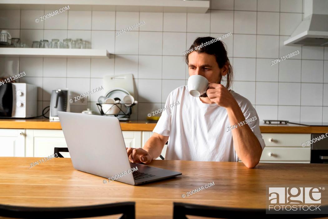 Stock Photo: Freelancer working from home on the kitchen and using laptop. Handsome smiling man holding cup of coffee and looking at laptop.