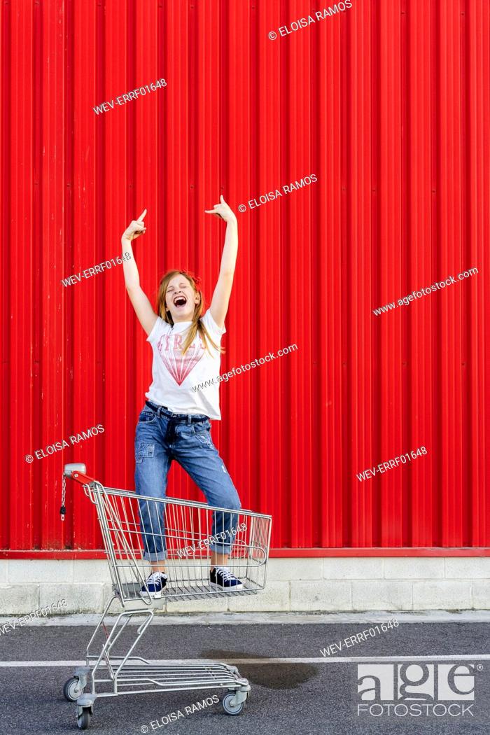 Stock Photo: Girl rocking in a shopping cart in front of red wall.
