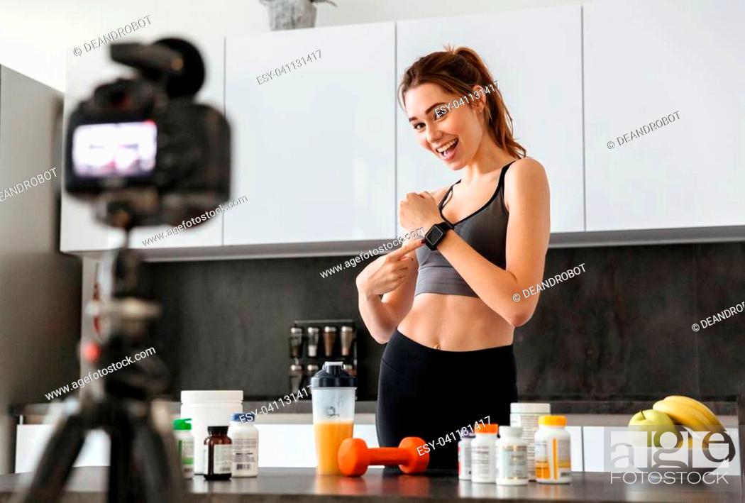 Stock Photo: Smiling healthy young girl recording her video blog episode about healthy food additives while standing at the kitchen at home and showing smart watch.