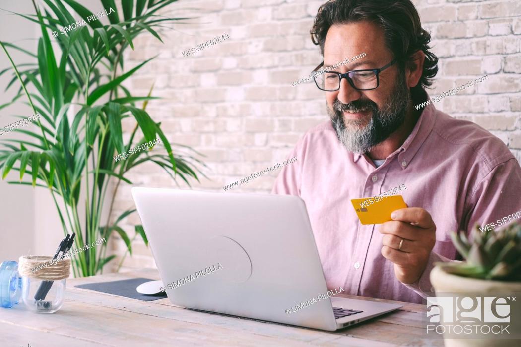 Stock Photo: Smiling businessman holding credit card doing online shopping through laptop at home.