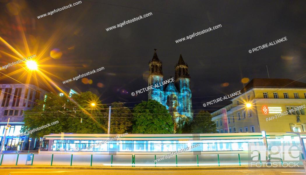 Stock Photo: 21 September 2021, Saxony-Anhalt, Magdeburg: A tram travels through the streets of the city center while the west building of Magdeburg Cathedral is illuminated.