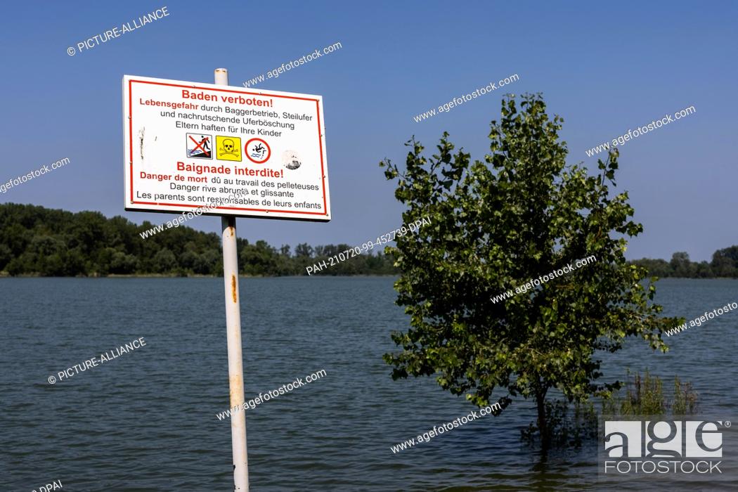 Stock Photo: 20 July 2021, Baden-Wuerttemberg, Neuried: A sign with the inscription ""Bathing prohibited! - Danger to life"" stands on the bank of a flooded area near the.