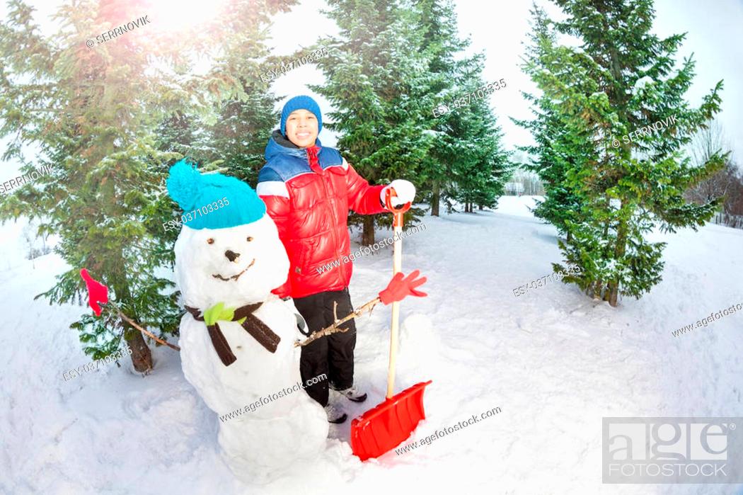 Stock Photo: Arabian boy with shovel standing near snowman in winter during day in the fir tree forest.