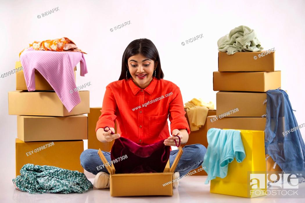 Stock Photo: A young woman unpacking new dress sitting amidst cardboard boxes, clothes and shoppingbags.