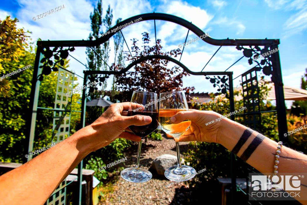 Stock Photo: couple hands toasting and cheering red and white wine glasses in the front of a winery garden gate, tasting restaurant patio in the background.
