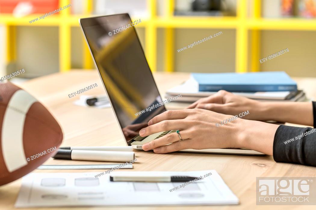 Stock Photo: Specialist is using a laptop on wooden table on the blurry background of the yellow shelves in the office. Next to her there are football ball.