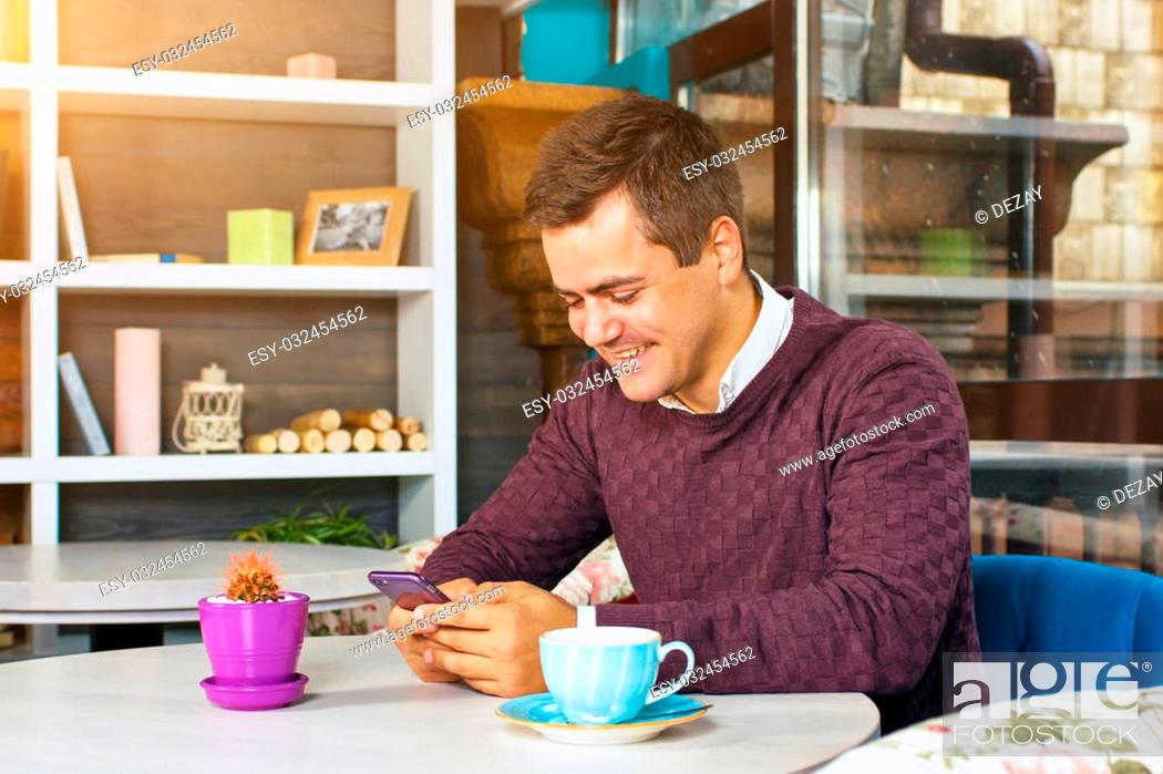 Stock Photo: Young man holding a mobile phone and smiling. Read the posts and drinking coffee.