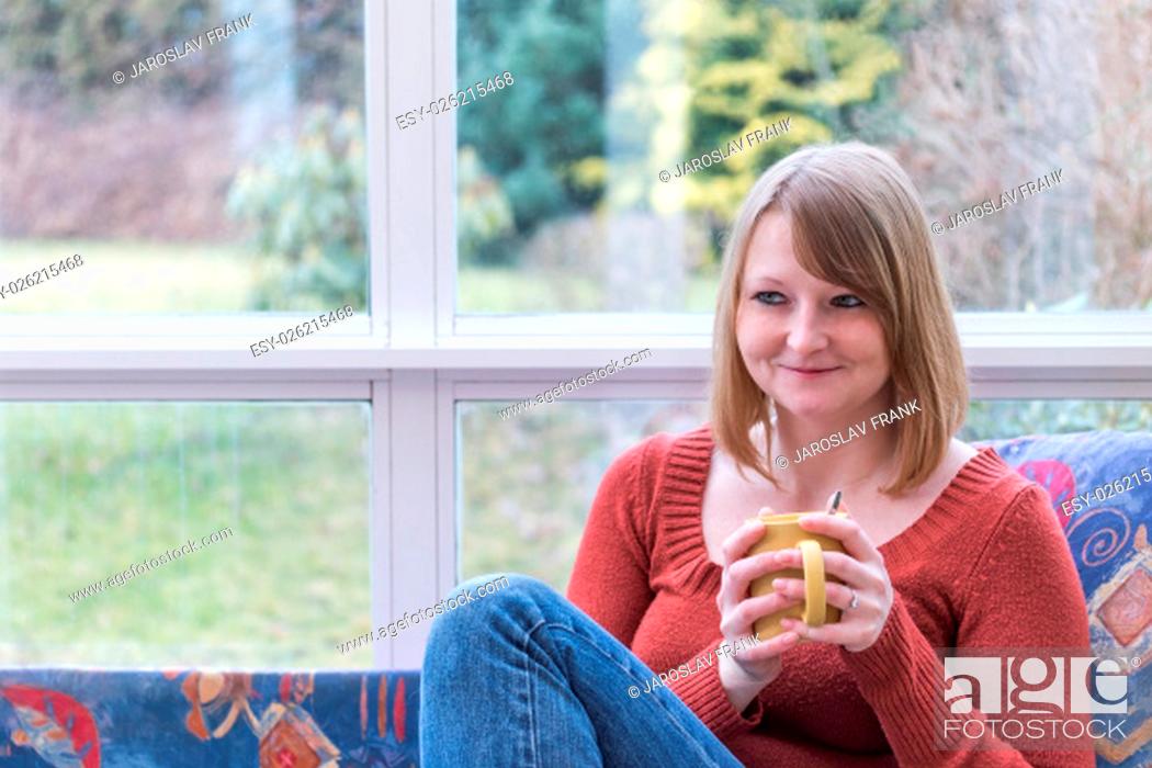 Stock Photo: Smiling young woman dressed in a brown sweater is sitting on the sofa in conservatory and she is looking to the right of the camera.