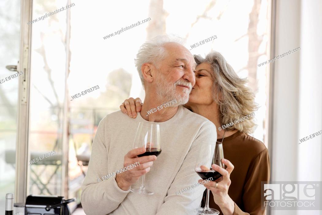 Stock Photo: Senior woman holding wineglass kissing man in front of window at hotel apartment.