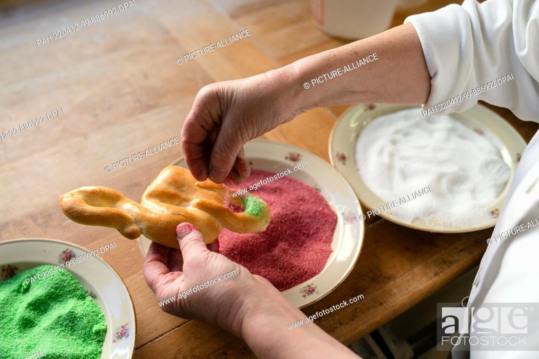 Stock Photo: PRODUCTION - 11 April 2022, Bavaria, Ostheim Vor Der Rhön: A woman sprinkles colored sugar on an ""Easter stork"" made from yeast dough.