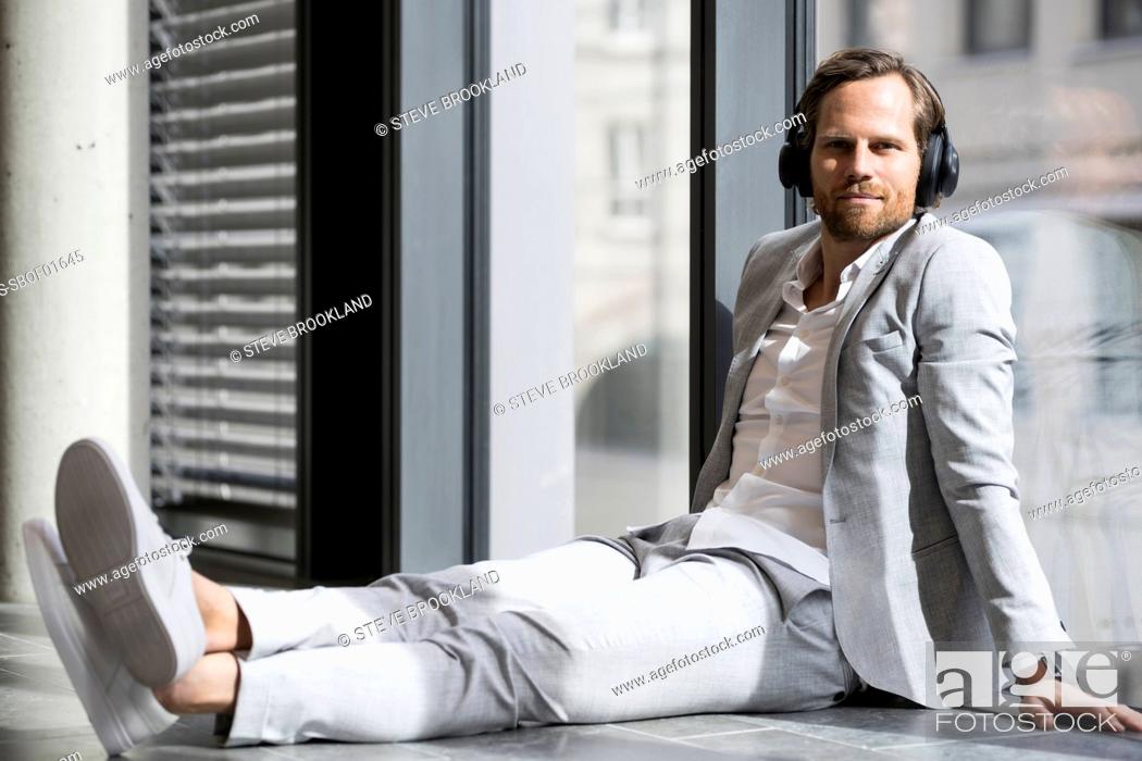 Stock Photo: Casual business man with headphones leaning onto window in modern office space listening to podcast.