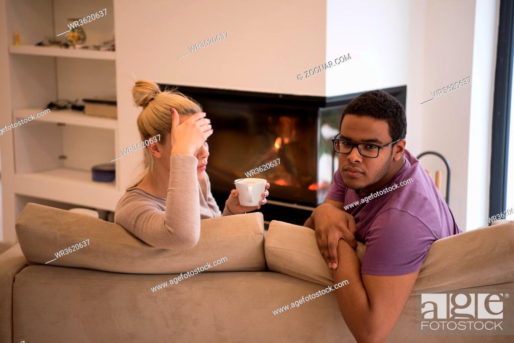 Stock Photo: Young romantic multiethnic couple sitting on sofa in front of fireplace at home, looking at each other, talking and drinking coffee.