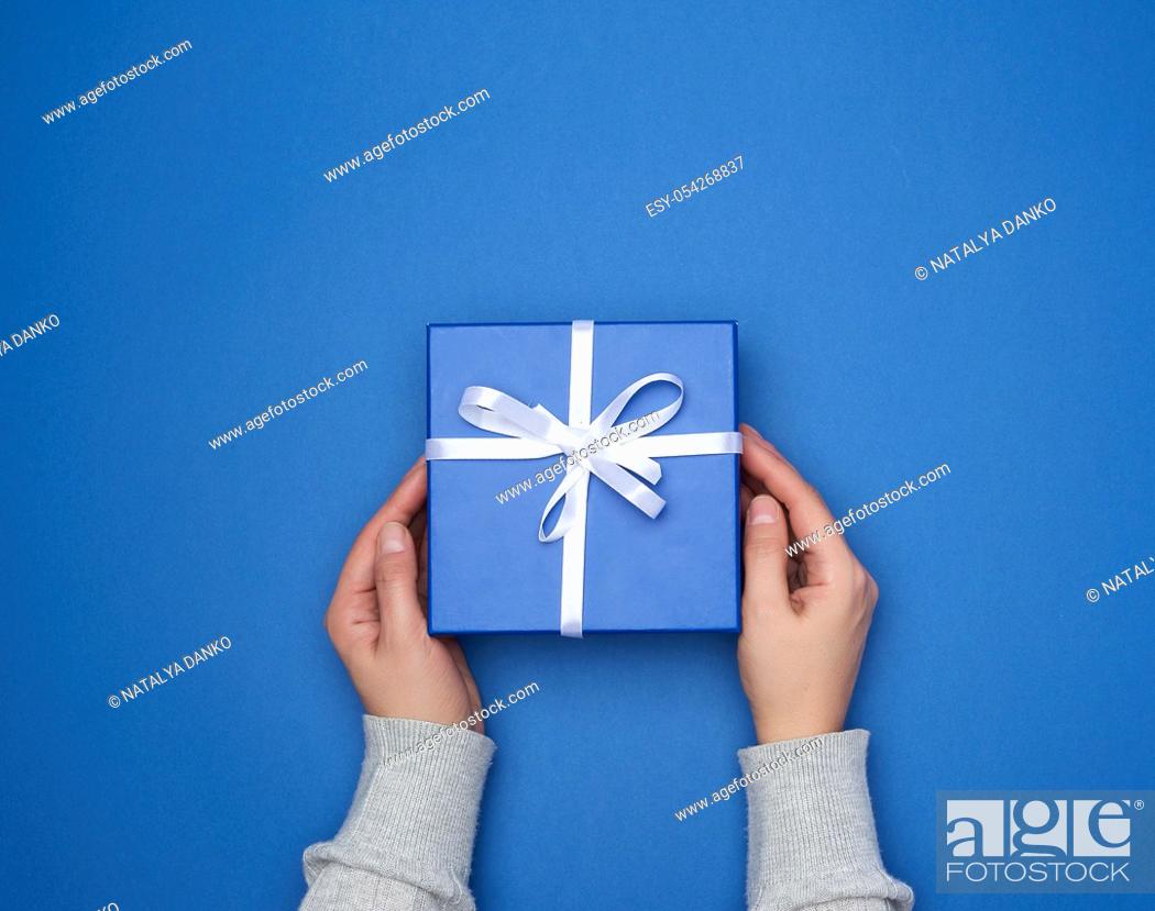 Stock Photo: two female hands in a sweater hold a square blue box with a bow on a blue classic background, top view, festive backdrop.