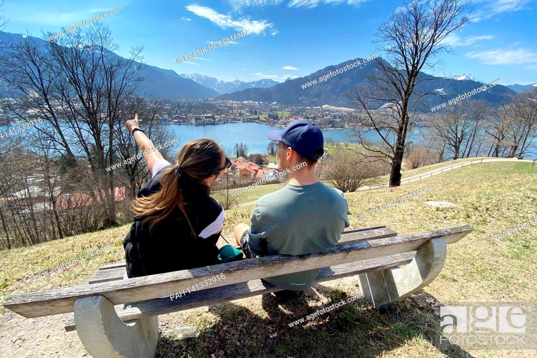 Stock Photo: Young couple take a rest on a bench. Hikers on the Hoehenweg over the Tegernsee with a view of Rottach Egern on April 1st, 2021.